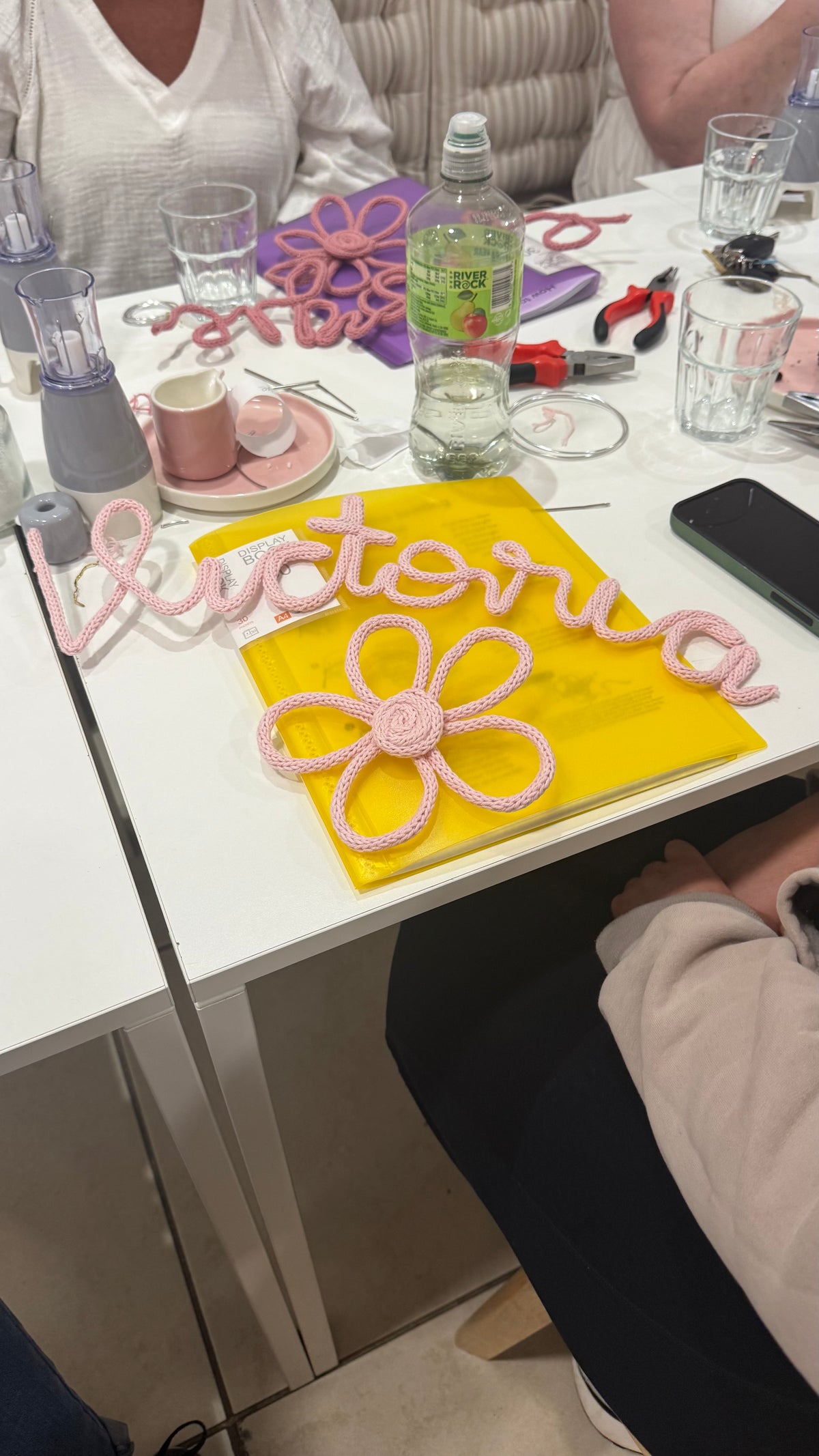 A white table at a craft workshop or class, featuring a pink crocheted script name 'Victoria' and a matching pink flower on a yellow backing board. The table is cluttered with various craft supplies, including wire, scissors, and glasses, and people are seated around it.