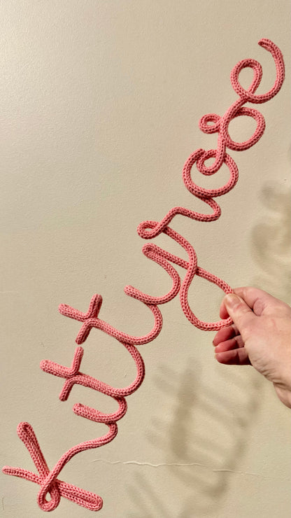 A salmon pink kitted wire wall sign spelling 'Kitty-Rose' being held by a hand against a beige wall.
