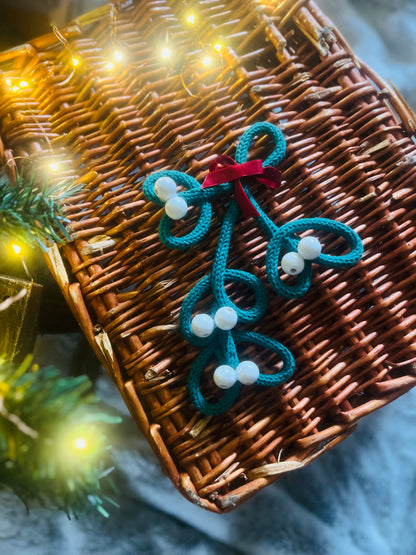 Handmade knitted wire mistletoe Christmas decoration with white wooden beads and burgundy velvet ribbon displayed on a wicker basket under a Christmas tree. 
